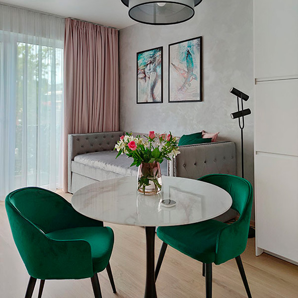 Moderní malometrážní byt s kulatým jídelním stolem, zelenými sametovými židlemi a světle šedou pohovkou, navržený pro krátkodobý pronájem v Praze.